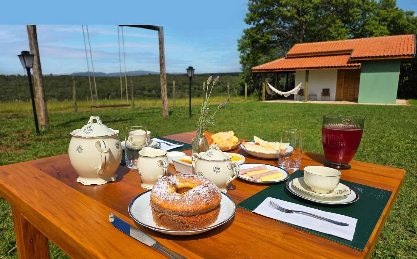 Mesa de café da manhã com bolo, bule e xícaras no gramado, diante de um chalé da Pousada Reserva Cambucá