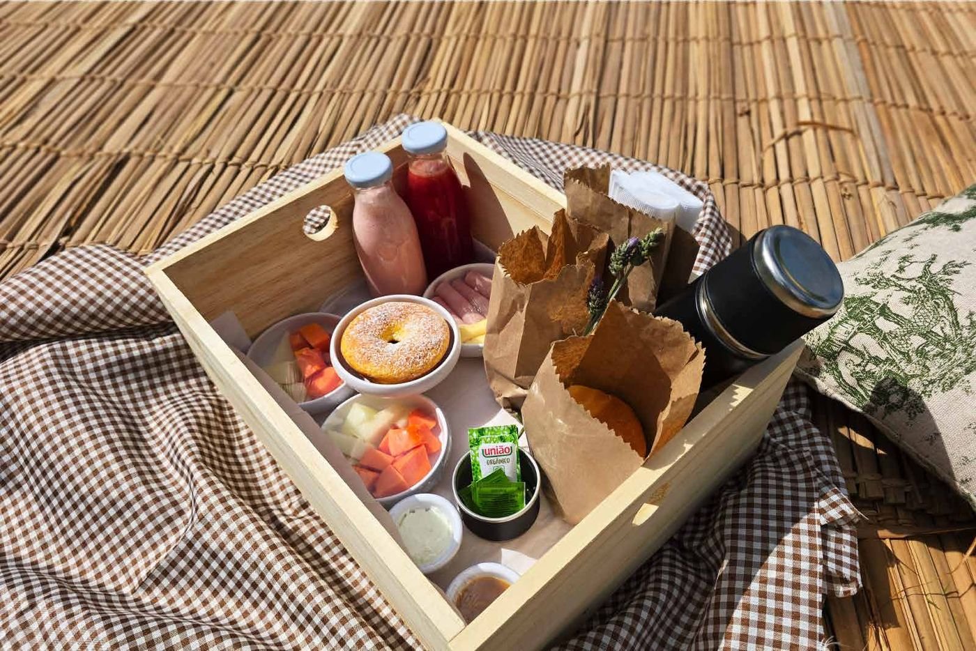 Cesta de madeira com café da manhã, sucos, pães e frutas sobre toalha xadrez