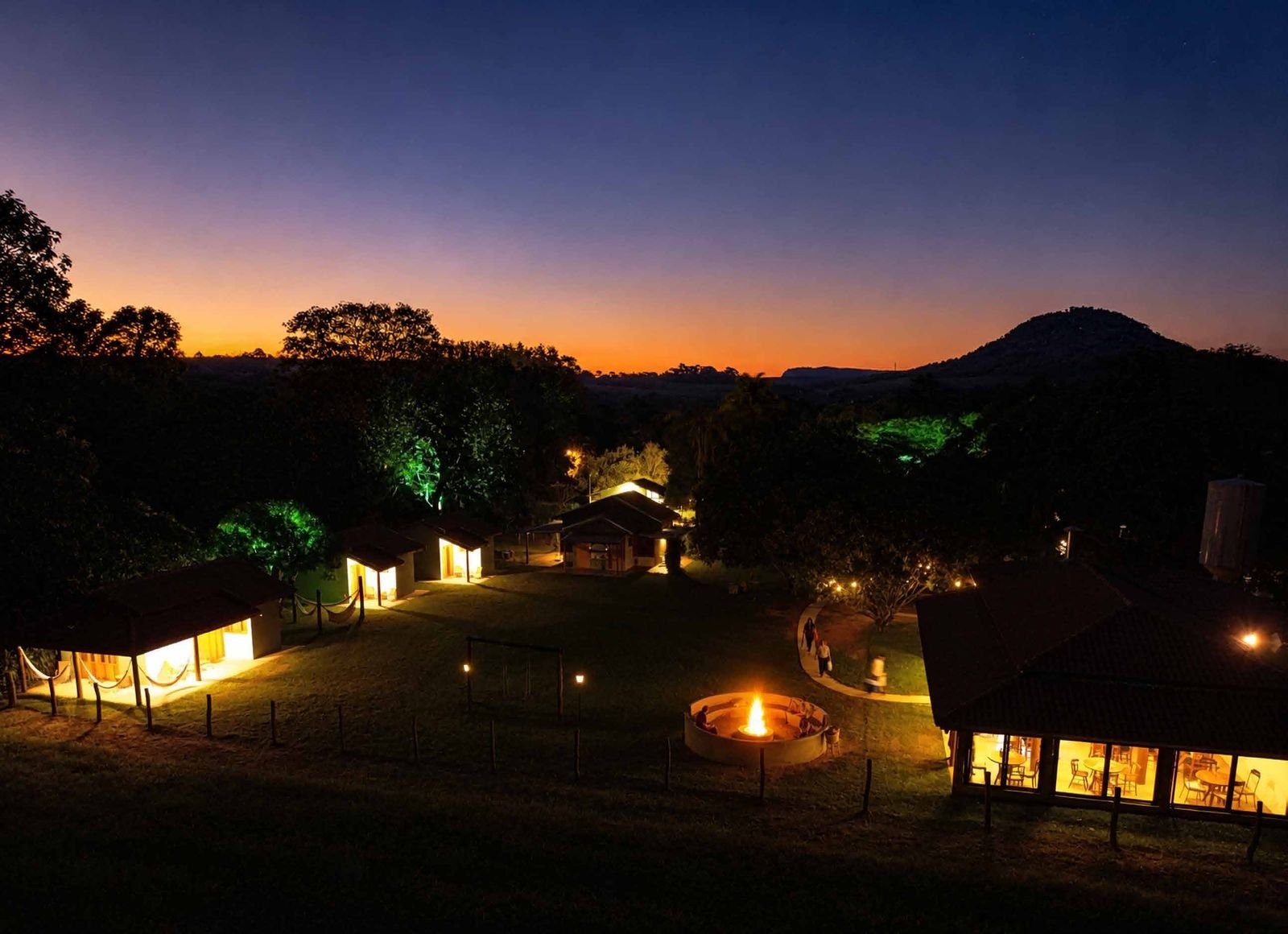 Pousada Reserva Cambucá ao anoitecer, com o fire pit aceso, o restaurante iluminado e a serra ao fundo