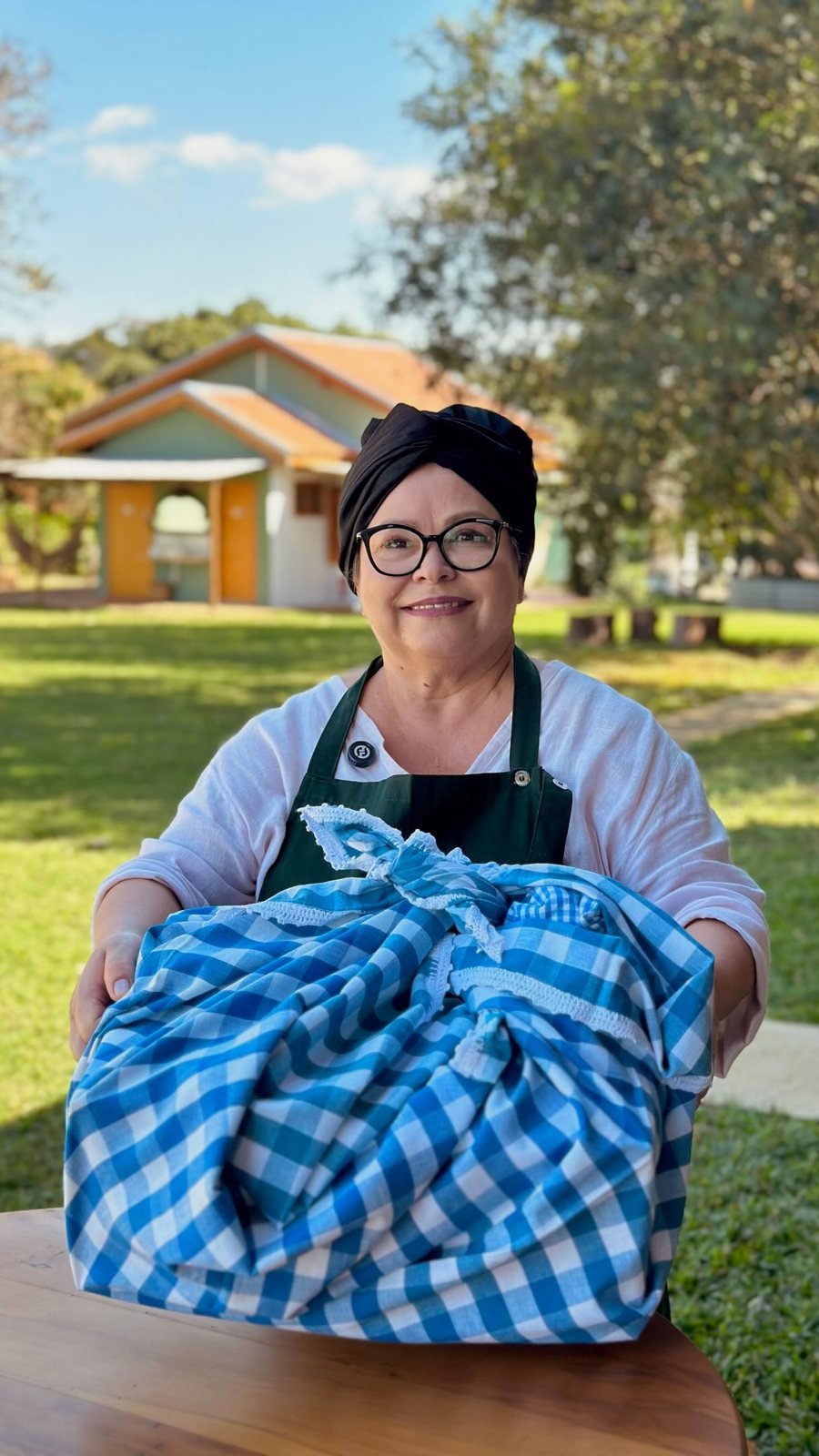 Sueli sorrindo com a cesta de café da manhã embrulhada em tecido xadrez azul, em frente aos chalés